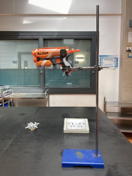 A photograph showing a Nerf gun mounted on a clamp stand pointing towards the a target