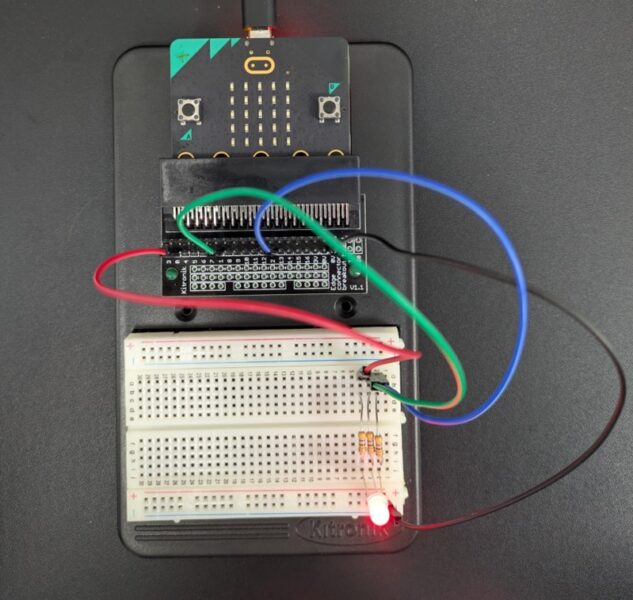 A microcontroller wired with 4 jumper cables in the colours green, red, blue and black to a breadboard where a red LED glows next to a resistor. 