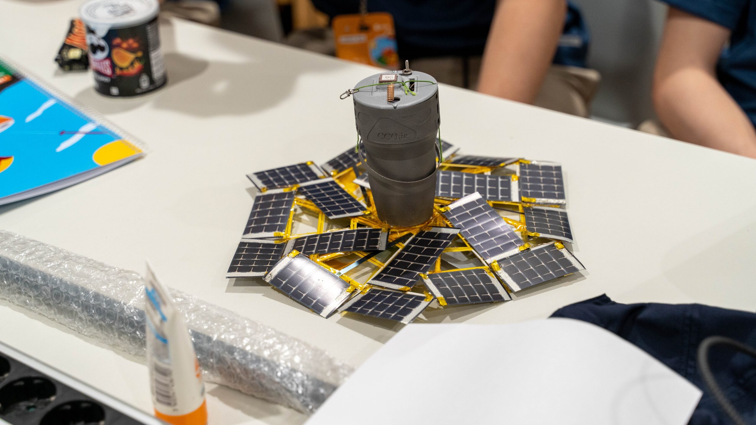A grey can-sized cylinder with some wires attached to it stands on a desk at the centre of a hexagonal array of credit-card-sized solar panels joined together with yellow plastic clips.