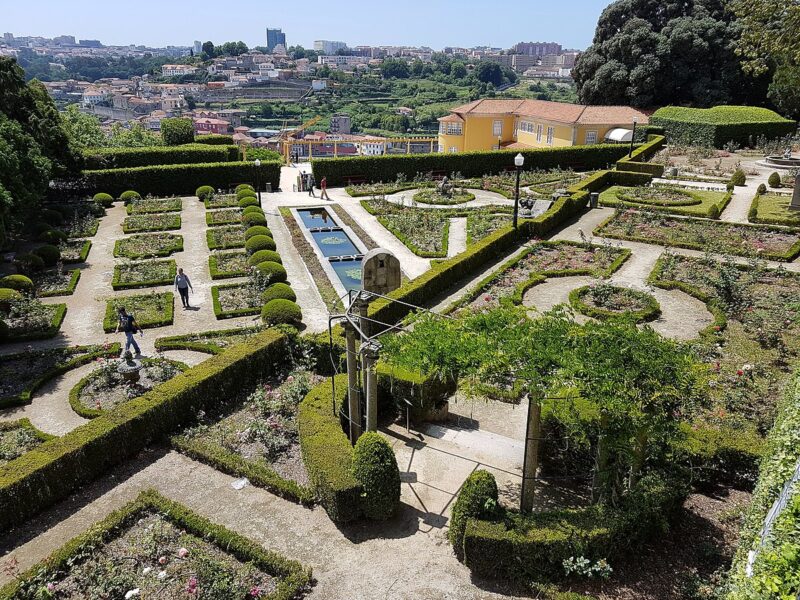 A photograph of a hillside garden with arrangements of geometric flower beds, square and circular, as well as a small rectangular pool. A panoramic city in the background.