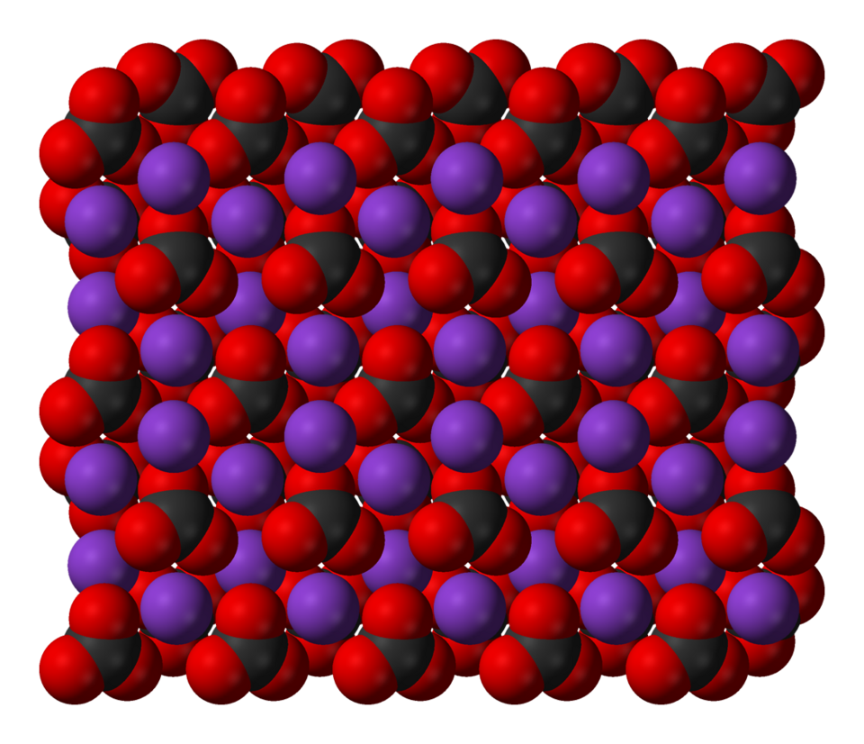 A 3D molecular structure of potassium carbonate. The carbonate ions are shown as black spheres surrounded by three red hemispheres and the potassium ions are shown as purple spheres. These two components are packed together in an alternating fashion to form a compact lattice.